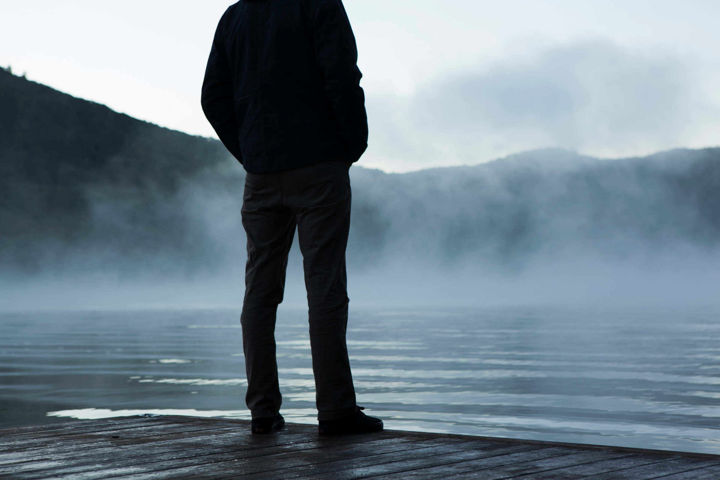 a person in shadow stands in front of a misty body of water surrounded by mountains